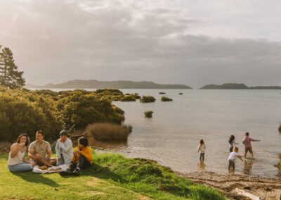 group of people sitting on grass by seashore having a picnic with children playing nearby in shallow water