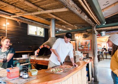 woman smiling and talking to 2 pizza chefs making pizza at a wood fired pizza oven