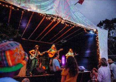 festivsl scene with musicians on stage below a home made canvas roof held up by bamboo poles, clourful crowd in front of stage