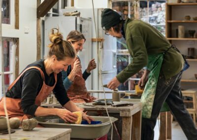 2 laughing women and one man all wearing aprons in a pottery workshop making pottery