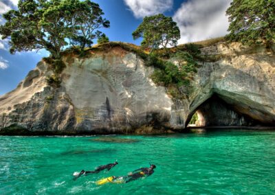 2 snorkelers in glassy clear green water swimming towards limestone cliffs topped with green trees and a blue sky